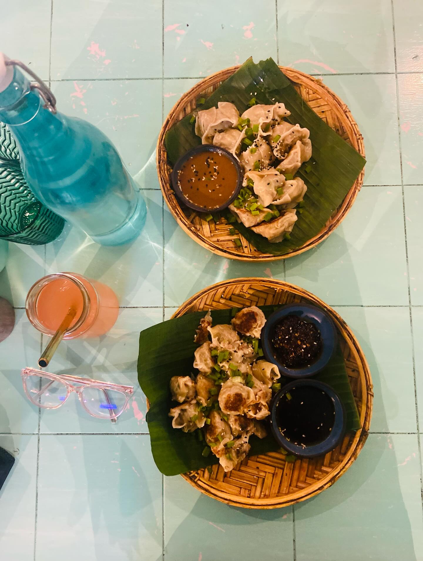 Two bamboo baskets of dumplings with dipping sauces, shot overhead on a teal tiled table